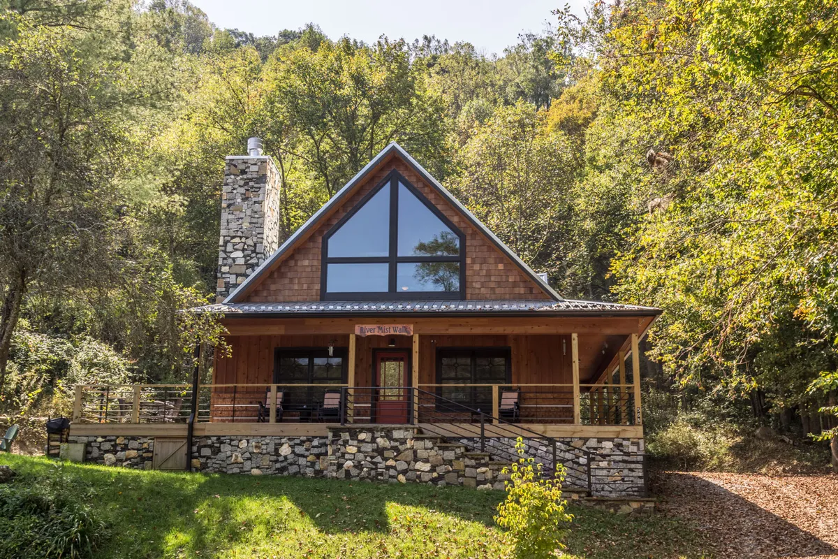 The exterior of a cozy cabin featuring large triangular windows, a stone chimney, and a welcoming wooden porch. The cabin is surrounded by lush trees and natural grounds, suggestive of a spacious, private setting perfect for outdoor exploration.