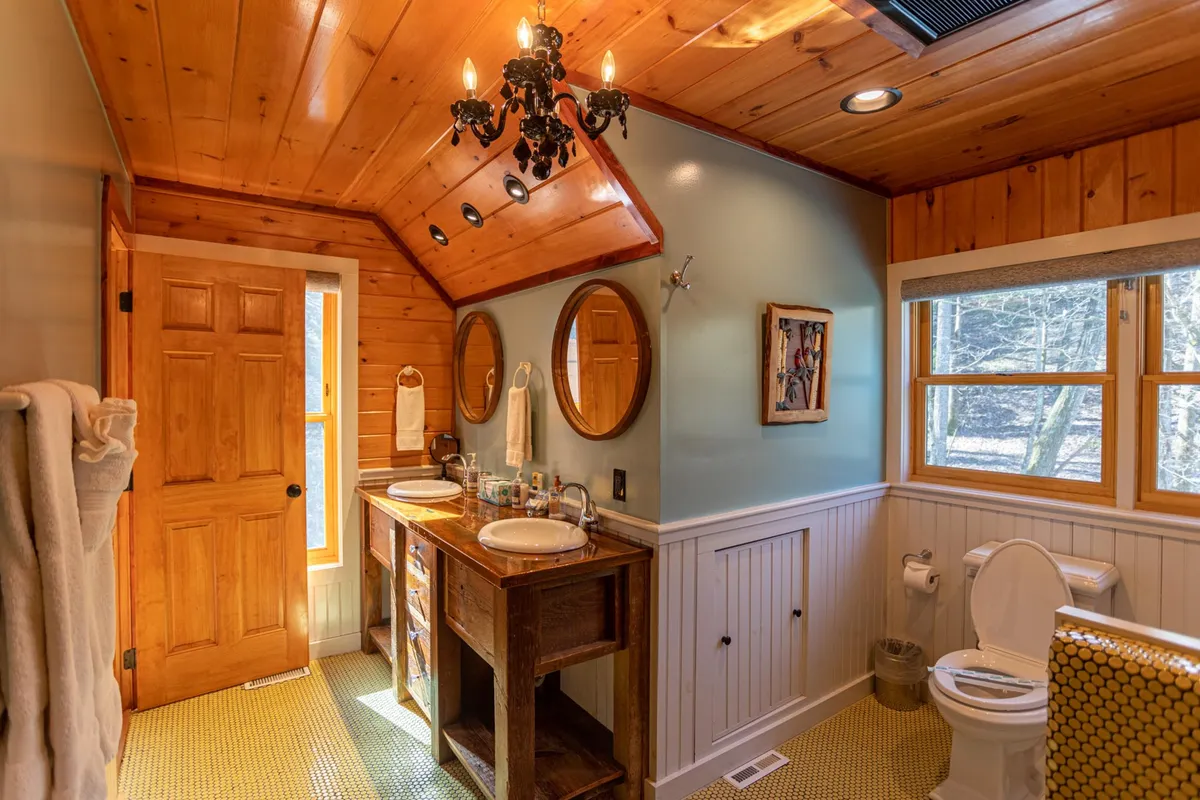 A serene bathroom view with a wooden double vanity, two circular mirrors, a chandelier, and abundant natural light streaming through a window. The vanity is crafted from warm-toned wood, enhancing the rustic yet elegant atmosphere.