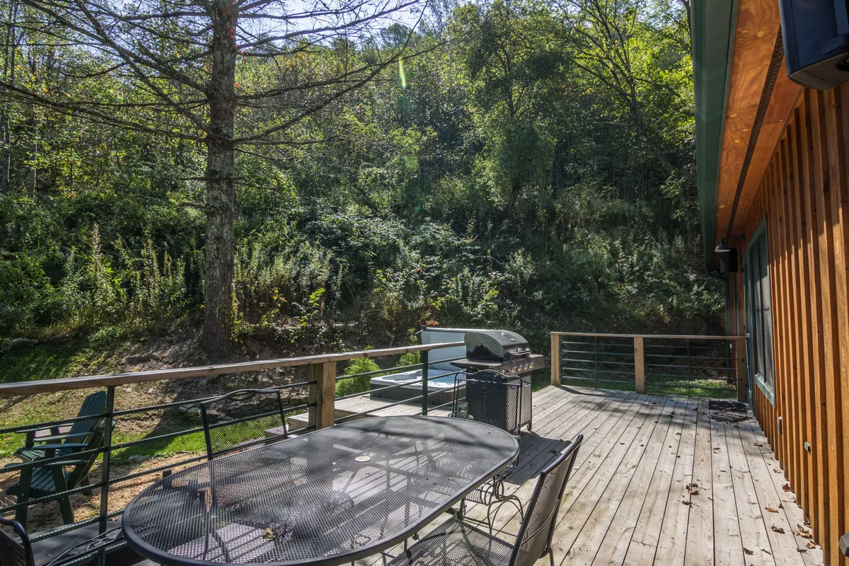 An exterior shot of a deck equipped with a dining table and grill, framed by lush green foliage and sturdy wooden railings. The inviting setup is designed for group dining, barbecuing, or simply relaxing while immersed in the quiet of the surrounding woods.