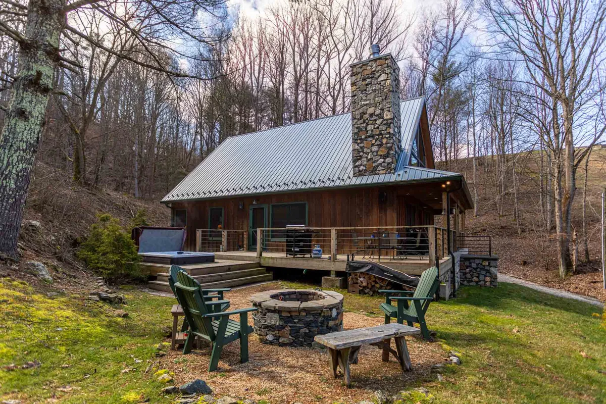 A cozy exterior view featuring a wooden deck with railing, visible fire pit, lawn chairs, and a welcoming cabin facade. The fire pit is ready for gatherings, and the deck is positioned for sunset watching and relaxation.