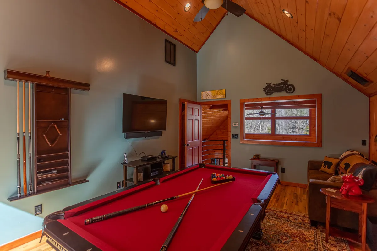 A casual, welcoming game room featuring a pool table, wall-mounted TV, wooden accents, and a leather couch. The doorway and room layout suggest a versatile space suited for both entertainment and lounging.