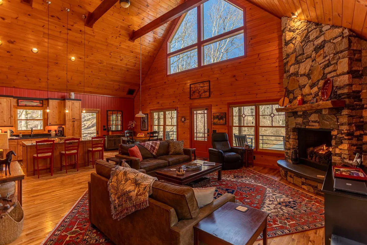The image presents a sweeping view of the great room, featuring a dramatic stone fireplace, vaulted wooden beams, large windows letting in abundant natural light, and a comfortable seating area that invites gathering. The dining space is visible, seamlessly connecting with the living area, showcasing the open-plan design.
