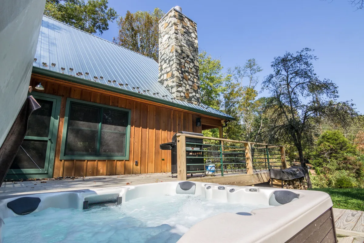 An inviting hot tub set on a spacious wooden deck, with a stone chimney adding rustic charm. The deck is surrounded by lush greenery, offering privacy and a direct connection to the natural mountain landscape. This outdoor amenity is perfect for relaxing or socializing while enjoying the fresh air and scenic views.