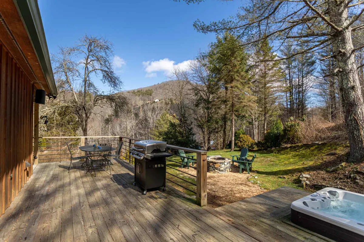 A wide-angle view of the main outdoor amenities: the deck with a grill, sunken hot tub, and a cozy fire pit surrounded by a tree line. The space is designed for barbecuing, relaxing soaks, and evenings around the fire.