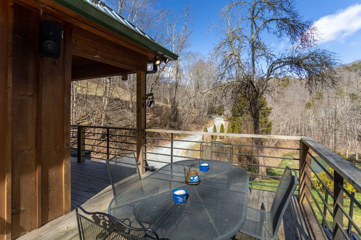 A broad wooden deck surrounded by vibrant trees, featuring a simple table set with blue cups, inviting guests to dine or relax outdoors. The deck offers a peaceful view of the natural landscape and leads to a gravel path, emphasizing the connection between indoor comfort and outdoor exploration.