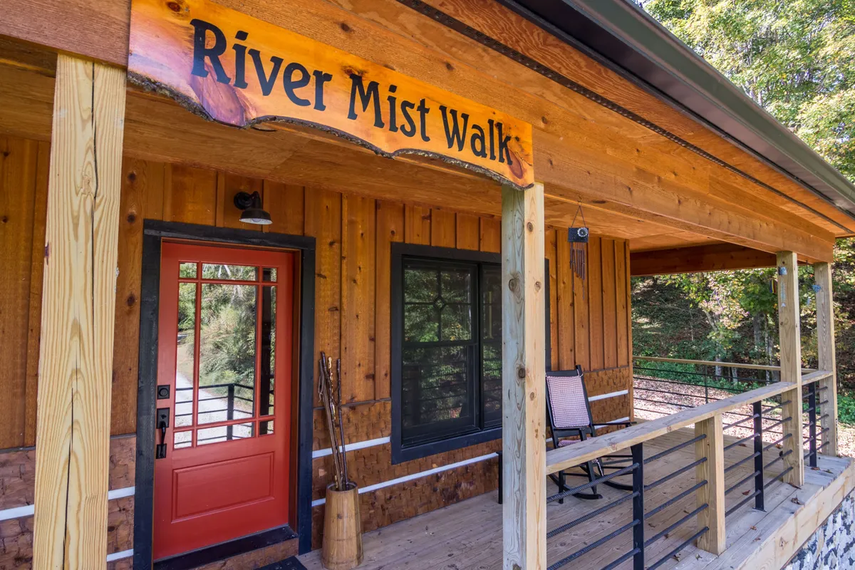 The image shows a welcoming wooden porch with a distinctive red door and a sign reading 'River Mist Walk.' A rocking chair sits on the porch, surrounded by lush greenery, creating an inviting entrance that hints at the property's connection to nature and the outdoors.