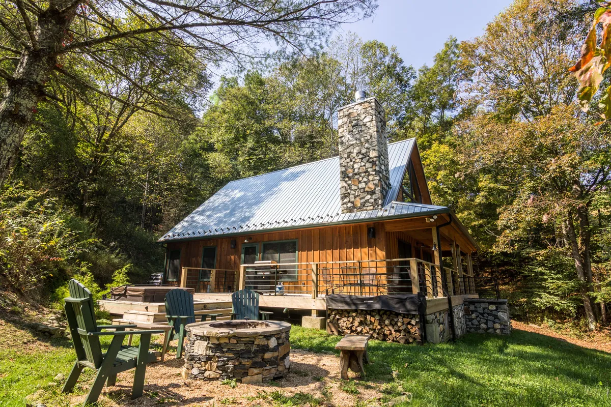A spacious wooden deck with a fire pit set among tall trees, stone chimney visible, and comfortable seating. The area feels peaceful and is ideal for relaxing after exploring, gathering around the fire, or enjoying sunset views amid nature.