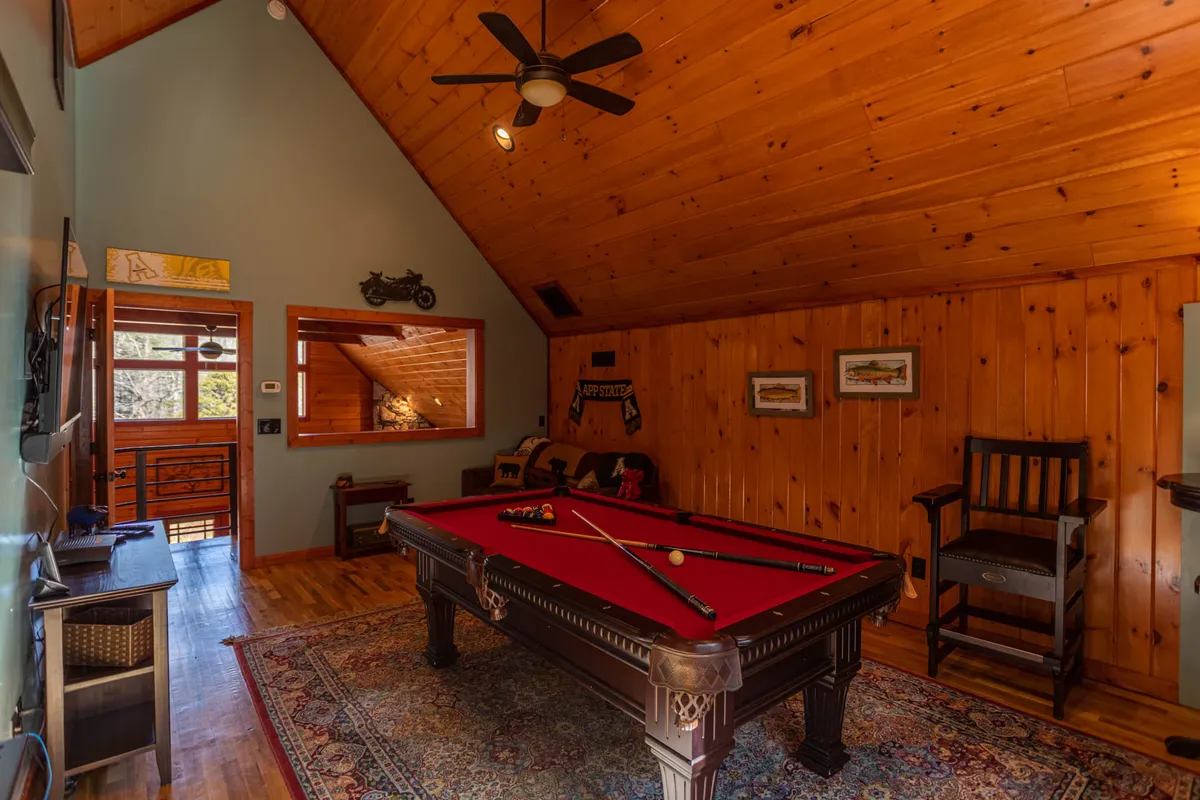 A warm, inviting game room with a central pool table, cozy wooden walls, a ceiling fan, and a comfortable seating area. A large decorative mirror and natural light contribute to a friendly, communal atmosphere.