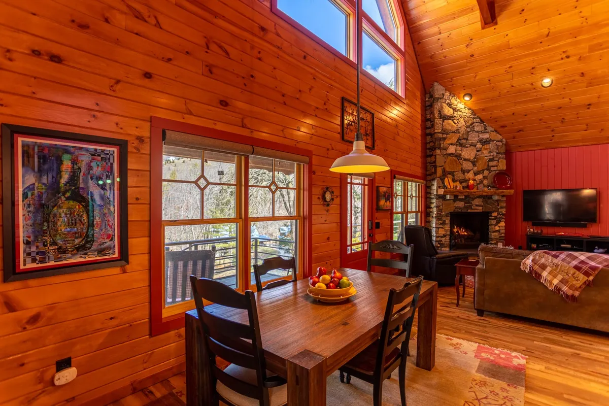 A warm, inviting interior shot focusing on the dining table adorned with a fruit bowl, the stone fireplace in the background, and expansive windows that frame the mountain views. The scene radiates an atmosphere of togetherness and comfort, with subtle touches of artwork and cozy lighting.